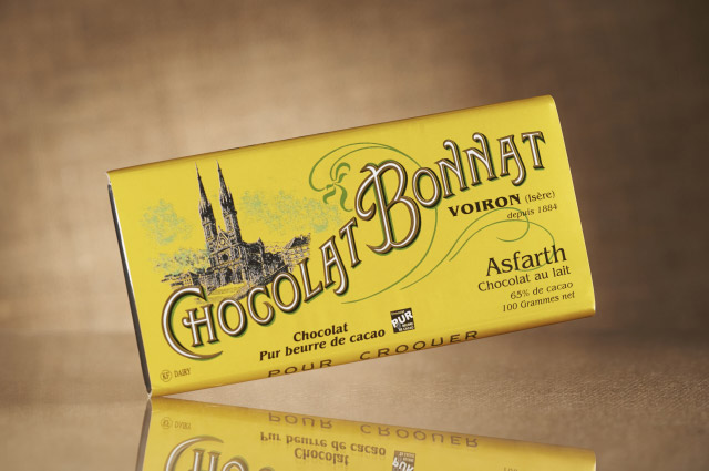 L’image de gauche présente une tablette de chocolat Bonnat 65% de cacao Grand cru Lait Asfarth, dans son emballage jaune, sur fond beige foncé.