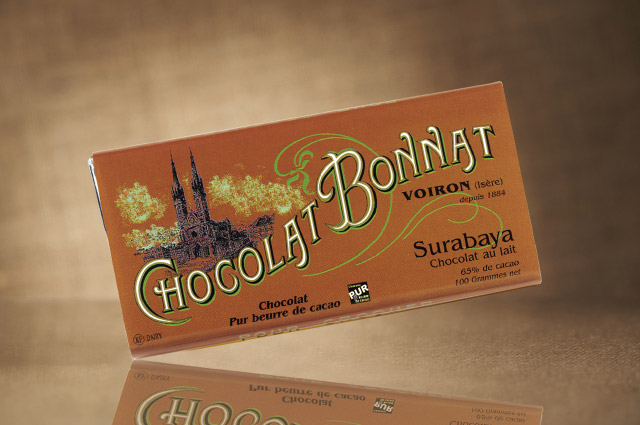 L’image de droite présente une tablette de chocolat Bonnat 65% de cacao Grand cru Lait Surabaya, dans son emballage marron clair, sur fond beige foncé.